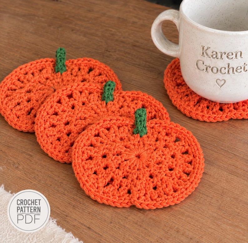 May include: Three orange pumpkin-shaped crochet coasters with green stems, arranged in a row. A white ceramic mug with the text Karen Crochet and a heart icon sits on a fourth matching coaster. A circular white label displays the text CROCHET PATTERN PDF.