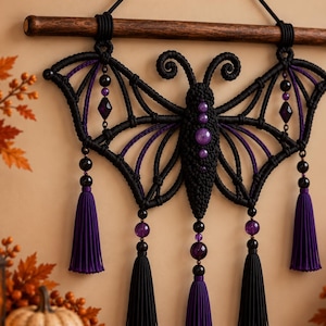 May include: A macrame wall hanging shaped like a butterfly with black cord, purple accents, and dark beads. The piece features intricate knotted wings, a central body with purple spheres, and five hanging tassels in black and purple colors.