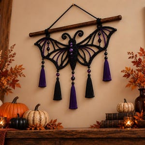 May include: A macrame wall hanging shaped like a bat with a black cord body and purple accents. The design features intricate knotted wings, a central body with purple beads, and five hanging tassels in alternating black and purple colors.