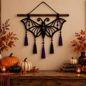 May include: A black macrame wall hanging shaped like a bat with purple accents and beads. The design features intricate knotted wings, a central body with purple beads, and five hanging tassels in alternating black and purple colors.