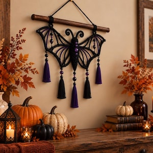 May include: A macrame wall hanging shaped like a bat with purple and black cord, featuring purple and black beads and five hanging tassels. The bat is suspended from a horizontal wooden dowel by a black cord, creating a symmetrical design.