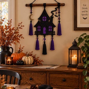 Könnte beinhalten: Ein schwarzer Makramee-Wandbehang in Hausform mit violetten Fenstern und einem Schornstein. Er besteht aus einem Holzstab, hängenden Perlen, einem goldenen Mondsichel-Anhänger, einer kleinen Fledermaus und fünf langen violetten und schwarzen Quasten.