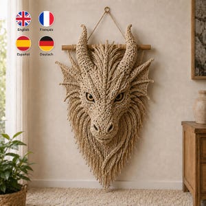 Op de afbeelding: Een beige macramé wandhanger in de vorm van een drakenkop met twee grote hoorns, puntige oren en een gestructureerde manen. Het stuk hangt aan een horizontale houten stok met een dun koord en bevat ingewikkeld knoopwerk en gele ogen.