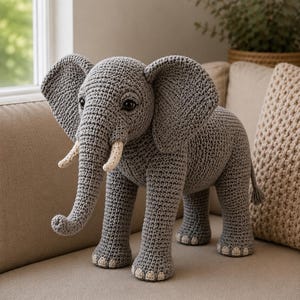 Puede incluir: Un juguete de elefante tejido a ganchillo en color gris con orejas grandes, una trompa y dos colmillos de color crema. El elefante tiene ojos de color marrón oscuro y detalles claros en los pies. El tejido crea un aspecto suave y detallado.