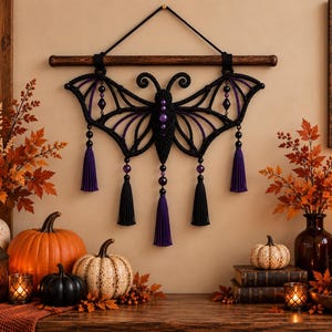 May include: A black macrame wall hanging shaped like a bat with purple accents and beads, suspended from a horizontal wooden rod. The design features symmetrical wings, a central body with purple beads, and five hanging tassels in black and purple.