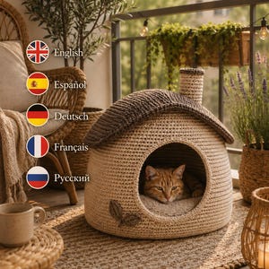 Pode incluir: Uma casa para gatos em crochê bege com um telhado castanho escuro e uma pequena chaminé. A casa apresenta uma entrada circular com uma almofada interior macia e uma aplicação decorativa em forma de folha na lateral. Um gato malhado descansa dentro.