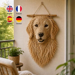 May include: A beige macrame wall hanging shaped like a golden retriever dog head. The piece features intricate knotted cord details, a realistic snout, dark eyes, and long fringe representing fur, suspended from a horizontal wooden dowel with a cord.