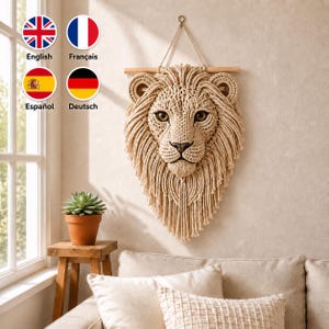 May include: A beige macrame wall hanging shaped like a lion head with a detailed mane, dark eyes, and a nose, suspended from a horizontal wooden dowel. The piece features intricate knotted cord patterns and long fringe detailing.