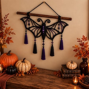 May include: A black macrame wall hanging shaped like a bat with purple accents, featuring a central body with purple beads, wing-like structures, and five hanging tassels in alternating black and purple colors, suspended from a dark wooden rod.