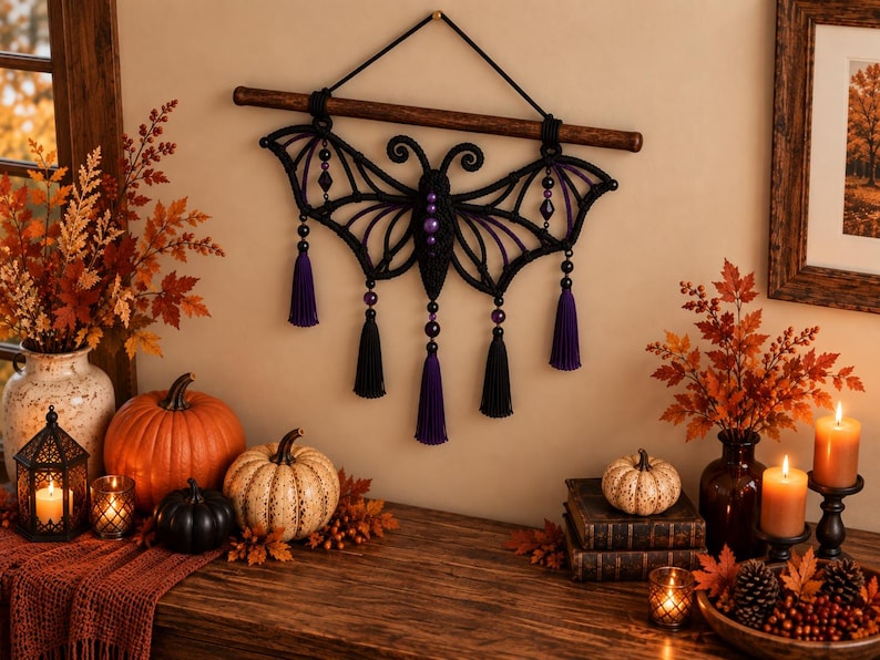 May include: A black macrame wall hanging shaped like a bat with purple accents, featuring a central body with purple beads, wing-like structures, and five hanging tassels in alternating black and purple colors, suspended from a dark wooden rod.