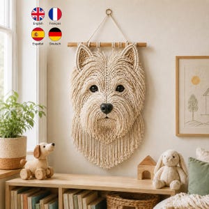 May include: A macrame wall hanging shaped like a West Highland White Terrier dog head, crafted from cream-colored cord. The piece features pointed ears, dark eyes, and a black nose, suspended from a horizontal wooden dowel with a hanging loop.
