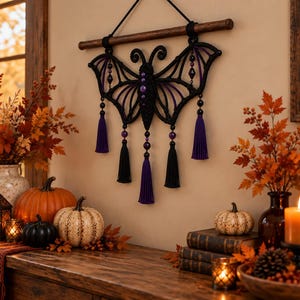May include: A black macrame wall hanging shaped like a bat with purple accents, featuring a central body with purple beads, two wings with geometric patterns, and five hanging tassels in alternating black and purple colors suspended from a wooden rod.