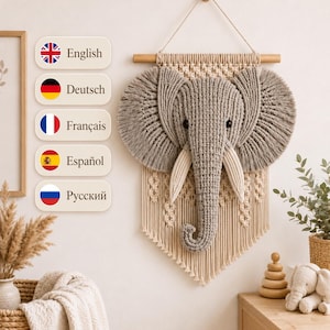 May include: A macrame wall hanging featuring a three-dimensional gray elephant head with large, fringed ears and cream-colored tusks. The elephant is centered on a cream-colored woven macrame panel suspended from a horizontal wooden dowel.