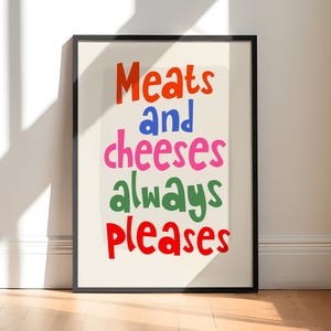 May include: A framed print with the words "Meats and cheeses always pleases" in colorful, playful lettering. The text is arranged in a stacked format, with each word in a different color, against a cream background.