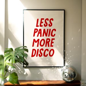 May include: A black framed print with the text "Less Panic More Disco" in red letters. The print is hanging on a white wall with a disco ball in the foreground.