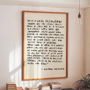 May include: A framed print with handwritten text by Anthony Bourdain. The text is in black ink on a white background, framed in a wooden frame. The print hangs on a white wall above a wooden cabinet with stacks of books.