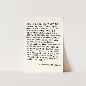 May include: A rectangular, off-white card with black handwritten text. The text is a quote by Anthony Bourdain, offering advice on life experiences, including dining and social interactions. The card is displayed against a plain background.