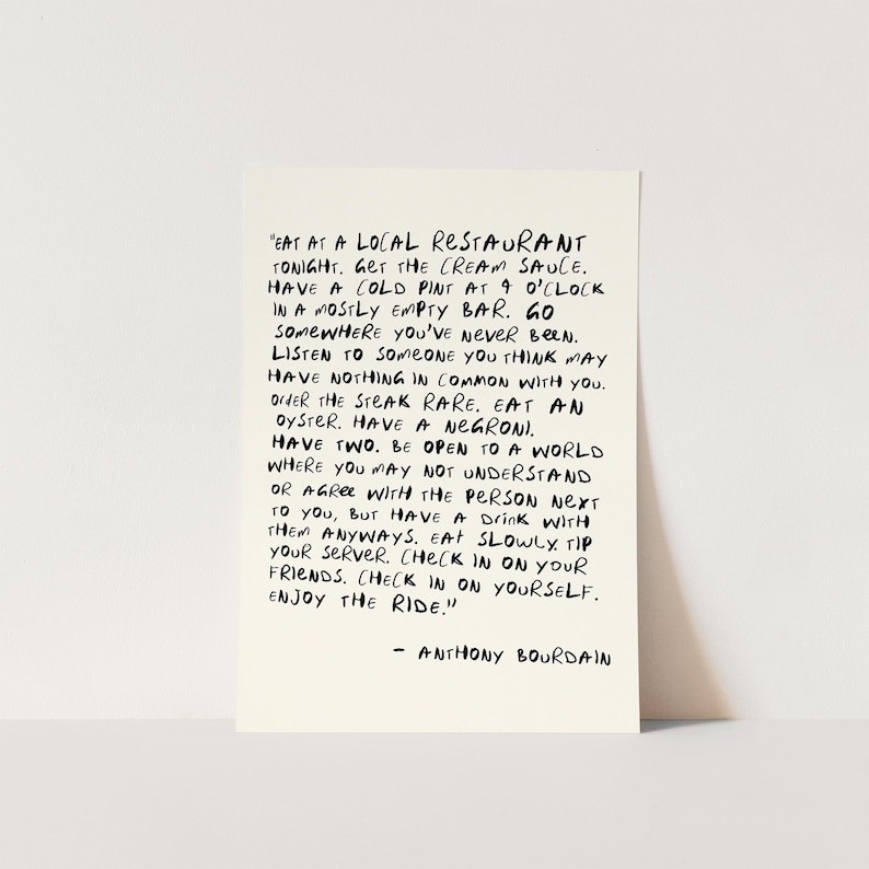 May include: A rectangular, off-white card with black handwritten text. The text is a quote by Anthony Bourdain, offering advice on life experiences, including dining and social interactions. The card is displayed against a plain background.