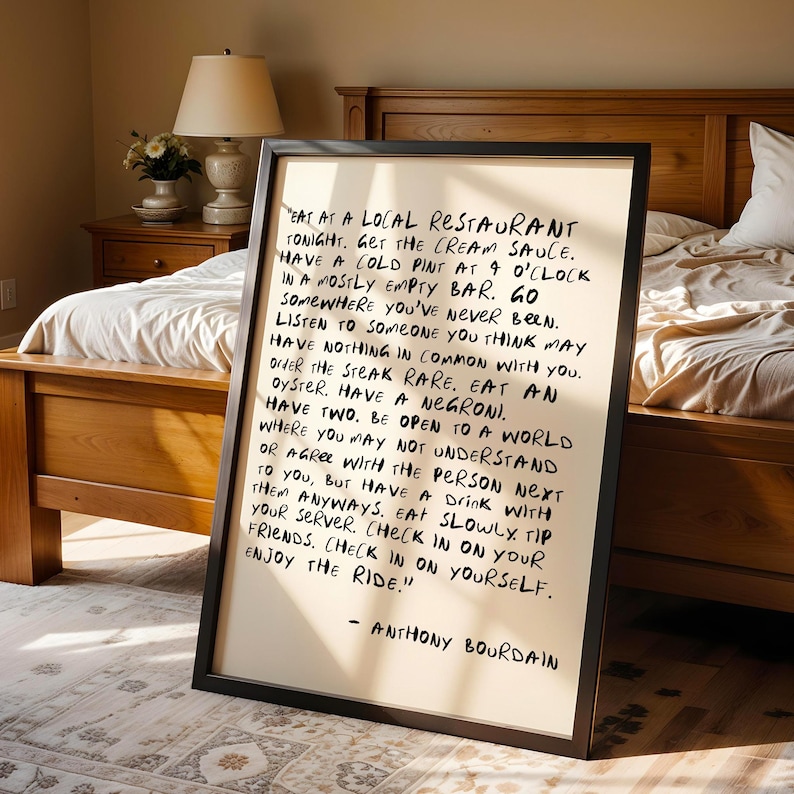 May include: A framed print with black lettering on an off-white background. The text, attributed to Anthony Bourdain, offers advice on dining and life, including "Eat at a local restaurant tonight." The frame is black.