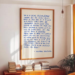 May include: A framed print with a wooden frame displays handwritten text in blue ink. The text, attributed to Anthony Bourdain, offers advice on life and enjoying experiences. The print hangs on a white wall above a wooden cabinet with books.