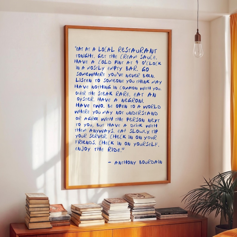 May include: A framed print with a wooden frame displays handwritten text in blue ink. The text, attributed to Anthony Bourdain, offers advice on life and enjoying experiences. The print hangs on a white wall above a wooden cabinet with books.