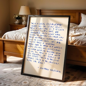 May include: A framed print with handwritten text in blue ink. The text, attributed to Anthony Bourdain, offers advice on life and enjoying experiences. The frame is black, and the print is displayed in a bedroom setting.