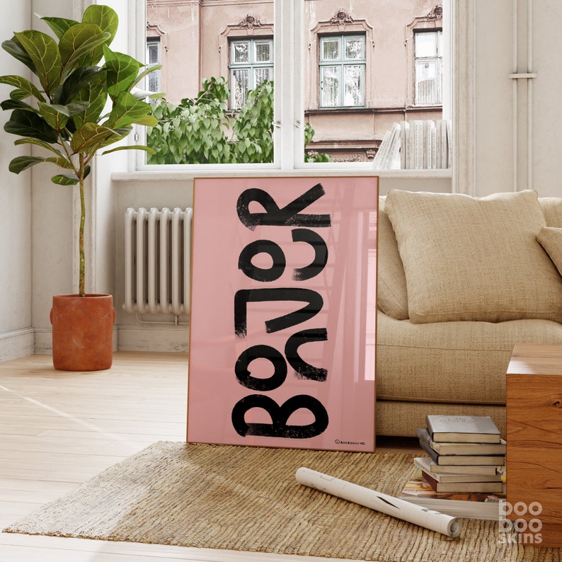May include: A pink framed poster with the word "BACK" written in black, stylised lettering. The poster is hanging on a wall in a room with a sofa and a plant.