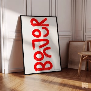 May include: A white framed poster with the word "BOOR" written in red, bold, and stylized lettering. The poster is leaning against a white wall.