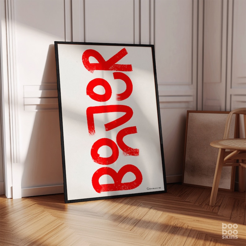 May include: A white framed poster with the word "BOOR" written in red, bold, and stylized lettering. The poster is leaning against a white wall.
