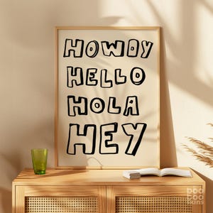 Peut inclure: Une affiche encadrée avec les mots "HOWDY, HELLO, HOLA, HEY" en lettres noires sur fond crème. L'affiche est dans un cadre en bois clair et est posée sur un meuble en bois avec un verre vert et un livre.