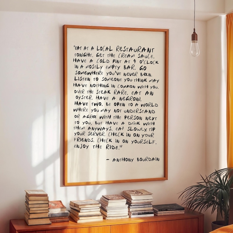 May include: A framed print with handwritten text by Anthony Bourdain. The text is in black ink on a white background, framed in a wooden frame. The print hangs on a white wall above a wooden cabinet with stacks of books.