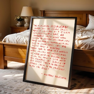 May include: A framed print with red handwritten text on a cream background. The text is a quote by Anthony Bourdain, offering advice on life and experiences. The frame is black, and the print is displayed in a bedroom setting.