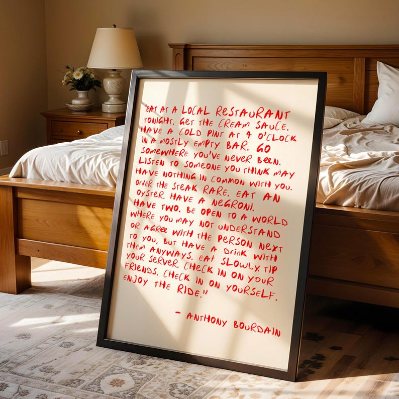 May include: A framed print with red handwritten text on a cream background. The text is a quote by Anthony Bourdain, offering advice on life and experiences. The frame is black, and the print is displayed in a bedroom setting.