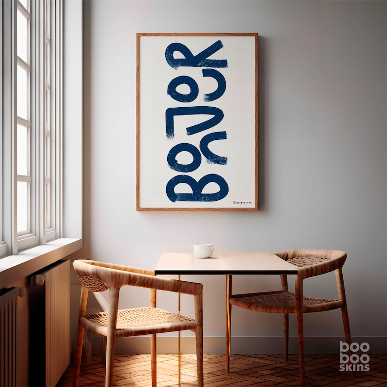 May include: A white framed poster with the word "BOJOK" written in large, blue, stylized letters. The poster is hanging on a white wall in a room with a wooden table and two chairs.