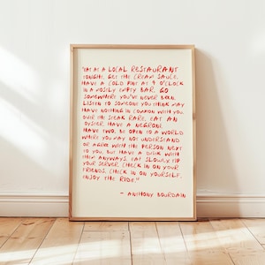 May include: A framed print with red handwritten text on an off-white background. The text is a quote by Anthony Bourdain, encouraging experiences like eating at a local restaurant and trying new things. The frame is a light brown color.