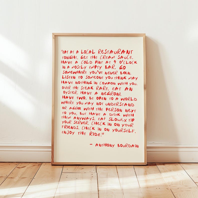 May include: A framed print with red handwritten text on an off-white background. The text is a quote by Anthony Bourdain, encouraging experiences like eating at a local restaurant and trying new things. The frame is a light brown color.
