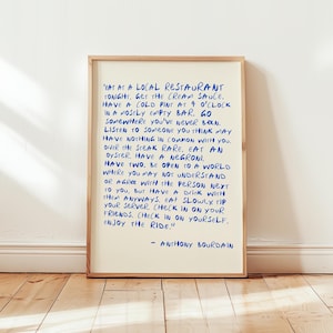 May include: A framed print with a cream-colored background and blue handwritten text. The text is a quote by Anthony Bourdain, offering advice on life and experiences. The frame is a light brown color, and the print is displayed on a wooden floor.