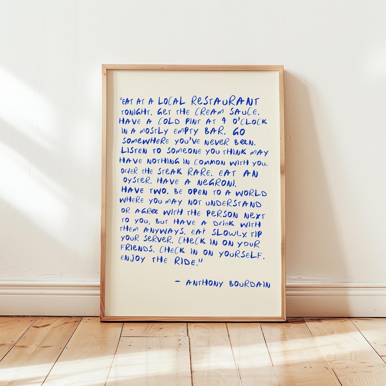 May include: A framed print with a cream-colored background and blue handwritten text. The text is a quote by Anthony Bourdain, offering advice on life and experiences. The frame is a light brown color, and the print is displayed on a wooden floor.