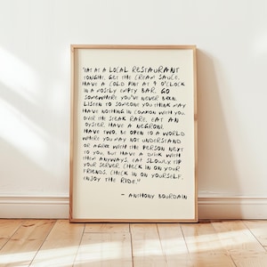 May include: A framed print with black handwritten text on an off-white background. The text is a quote by Anthony Bourdain, encouraging the reader to embrace new experiences and enjoy life. The frame is a light brown color.