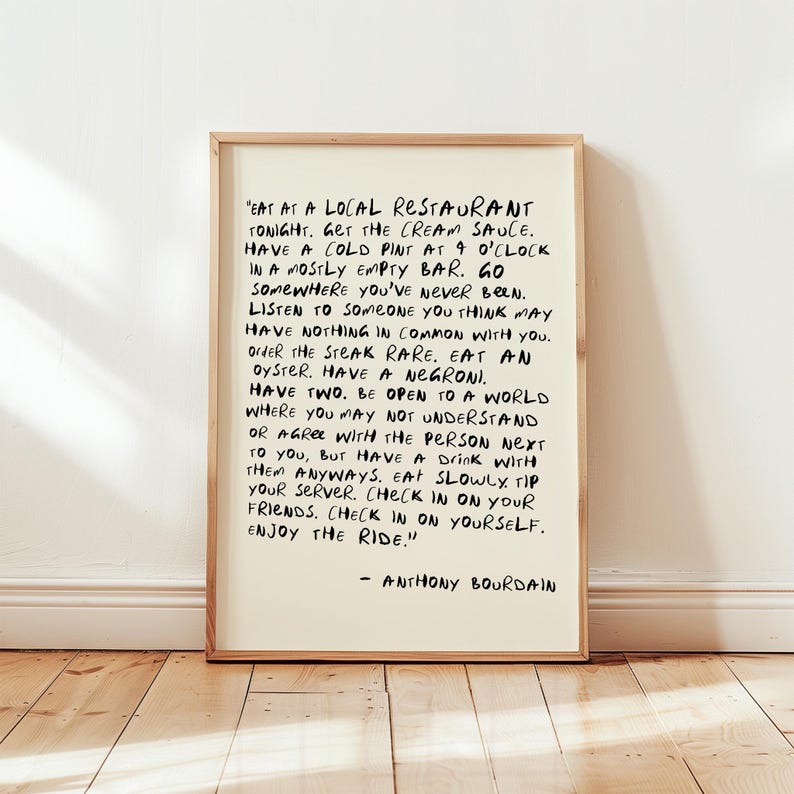 May include: A framed print with black handwritten text on an off-white background. The text is a quote by Anthony Bourdain, encouraging the reader to embrace new experiences and enjoy life. The frame is a light brown color.