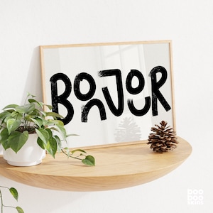 May include: A framed print featuring the word "Bonjour" in bold, black, stylised lettering. The print is hanging on a white wall above a wooden shelf with a pine cone on it.