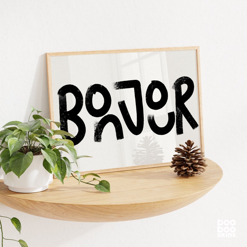 May include: A framed print featuring the word "Bonjour" in bold, black, stylised lettering. The print is hanging on a white wall above a wooden shelf with a pine cone on it.