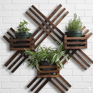 May include: A wooden wall-mounted planter with three dark brown wooden boxes, each holding a green potted plant. The boxes are set within a crisscross wooden frame against a white brick wall.