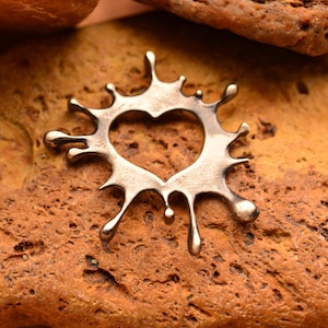 May include: A silver-colored pendant in the shape of a heart within a splash design. The pendant has a heart-shaped cutout in the center and droplet-like extensions around the edges. The pendant is displayed on a textured, reddish-brown surface.