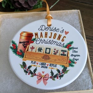 May include: A round white ceramic ornament with a gold ribbon, featuring a graphic of a Mahjong tile rack, a coffee cup labeled Mahjong Girl, pine branches, holly, and a pink bow. Text reads Denise's MAHJONG Christmas, Good Tiles Brighter Days, and PLAY LAUGH REPEAT 2026.