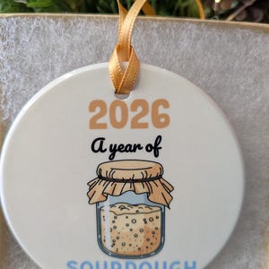 May include: A round white ceramic ornament featuring the text 2026, A year of, and SOURDOUGH in blue. The design includes a graphic of a glass jar filled with bubbly sourdough starter, topped with a tan cloth cover and tied with a gold ribbon.