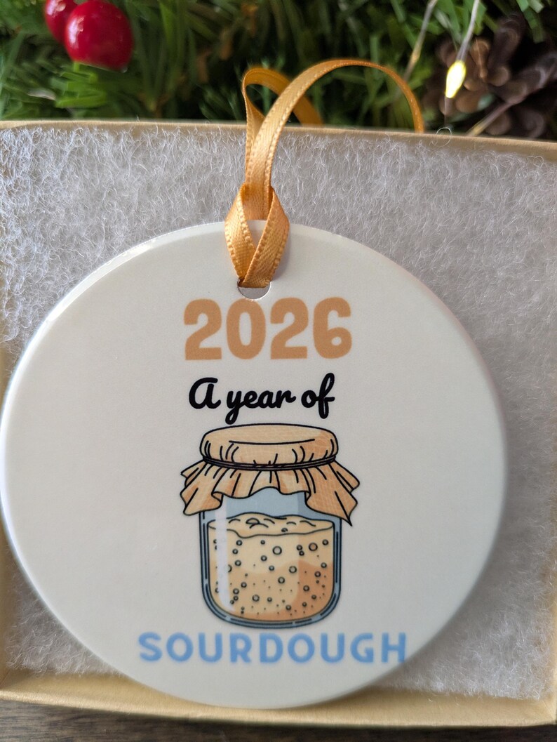May include: A round white ceramic ornament featuring the text 2026, A year of, and SOURDOUGH in blue. The design includes a graphic of a glass jar filled with bubbly sourdough starter, topped with a tan cloth cover and tied with a gold ribbon.