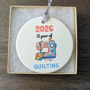 May include: A round white ceramic ornament with a grey ribbon, featuring the text 2026 A year of QUILTING and a colorful illustration of a sewing machine with a patchwork quilt piece, presented inside a square cardboard gift box.
