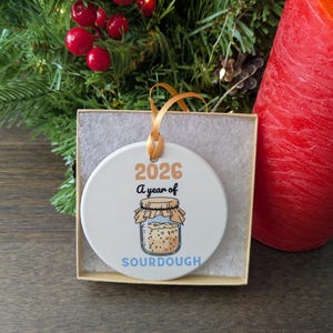 May include: A round white ceramic ornament with a gold ribbon, featuring the text 2026 A year of SOURDOUGH and a graphic of a glass jar filled with sourdough starter. The ornament is presented inside a square cardboard box with white padding.