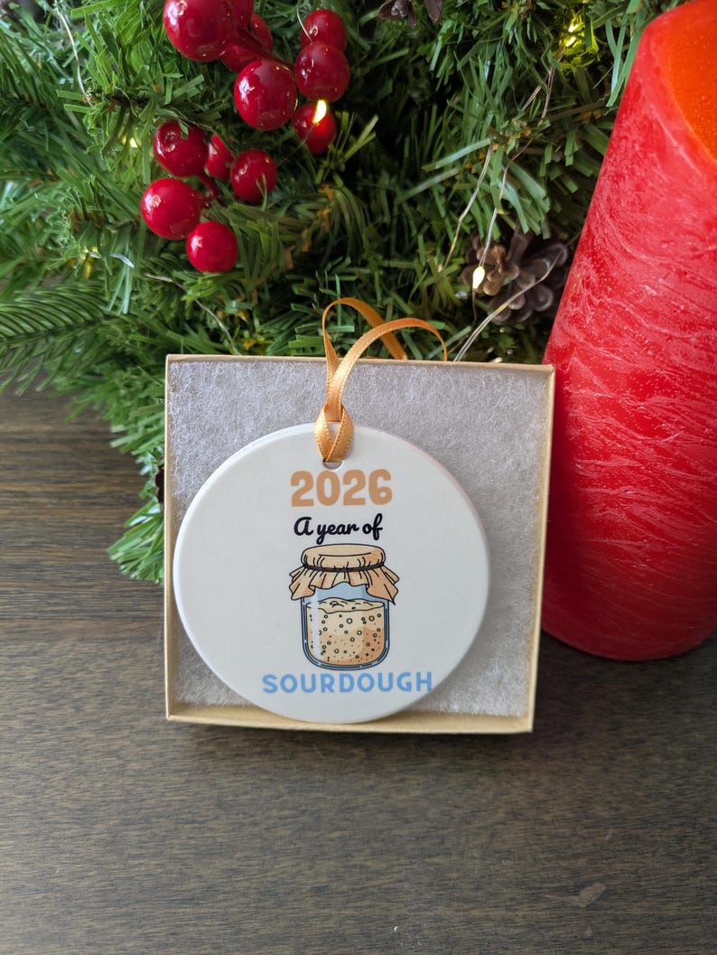 May include: A round white ceramic ornament with a gold ribbon, featuring the text 2026 A year of SOURDOUGH and a graphic of a glass jar filled with sourdough starter. The ornament is presented inside a square cardboard box with white padding.